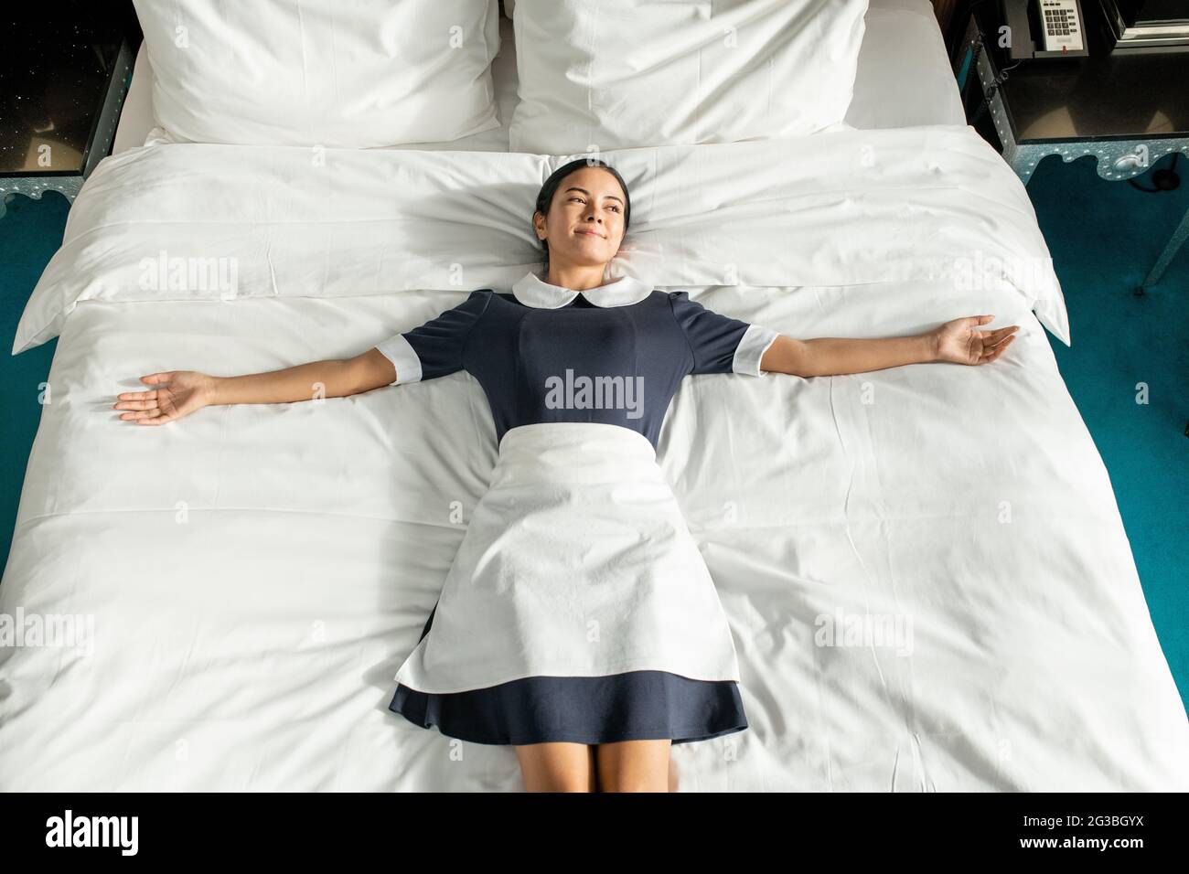 Fröhliche junge Zimmermädchen in Uniform, die auf einem großen Doppelbett liegt Stockfoto