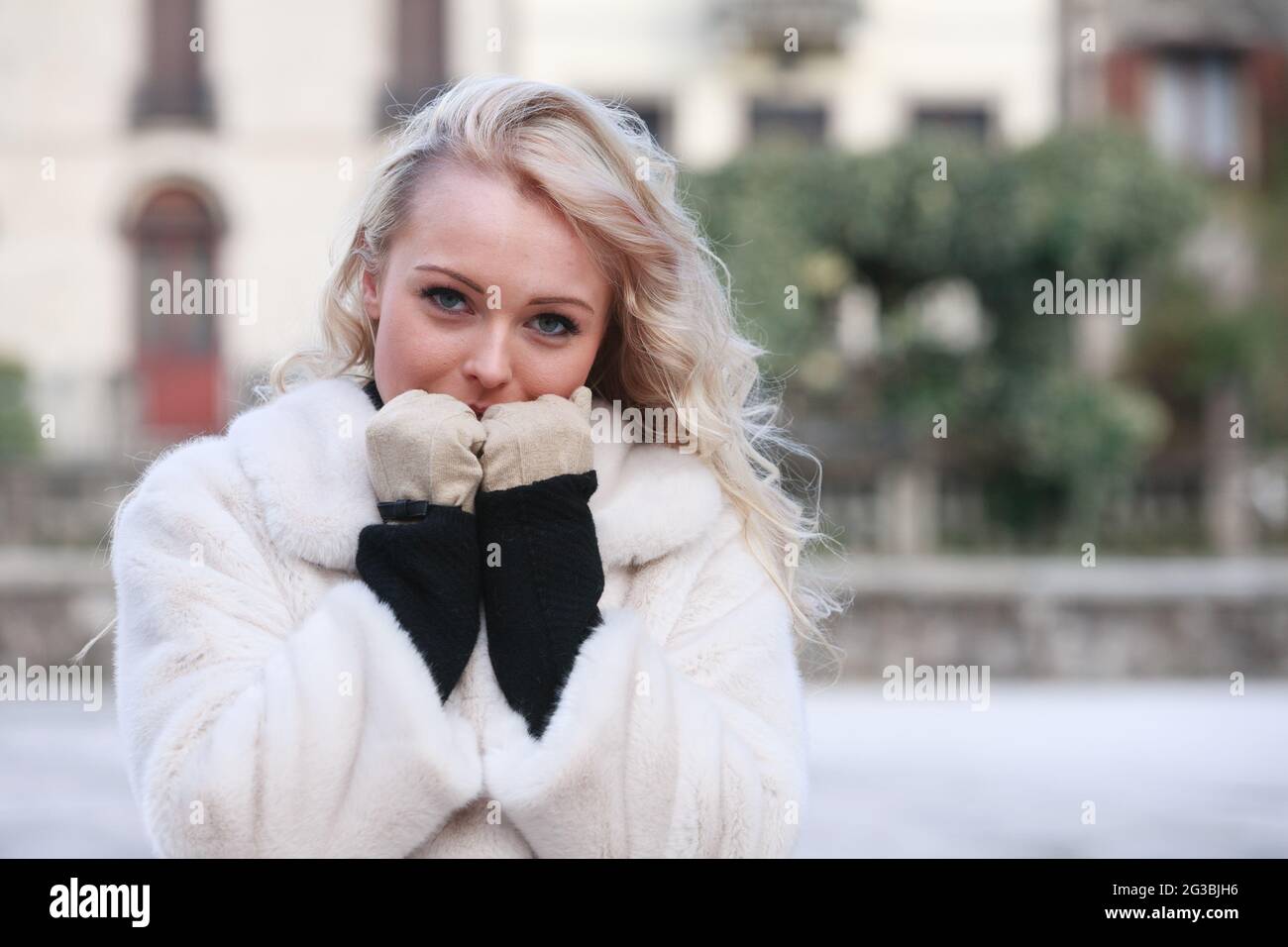 Junge blonde Frau in stilvollem, warmem Mantel, die an einem kalten Wintertag draußen steht und ihre Handschuhen an ihrem Gesicht hält, um sich warm zu halten Stockfoto