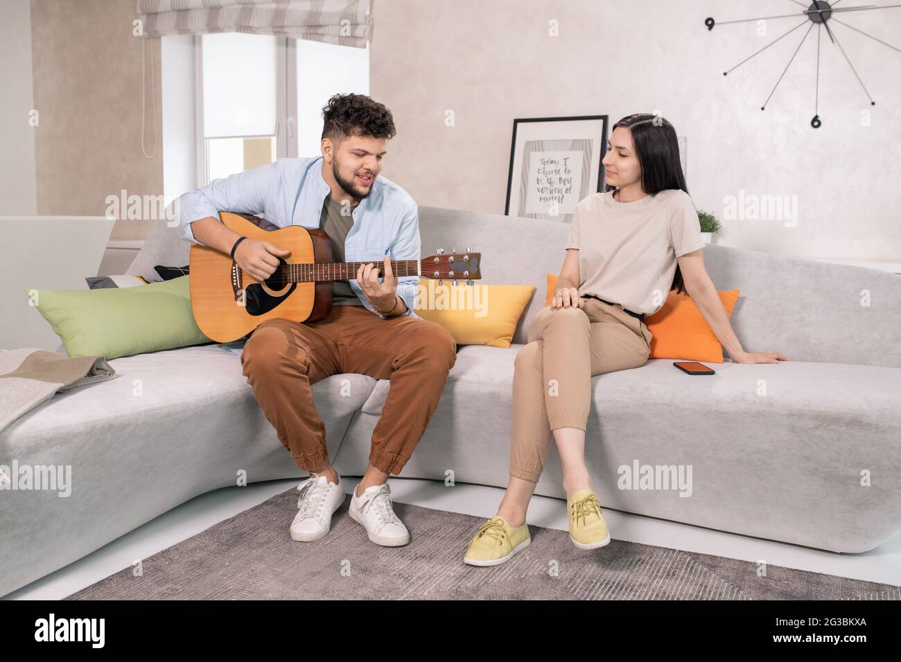 Junge Frau, die ihrem Mann zuhört, der in häuslicher Umgebung Gitarre spielt Stockfoto