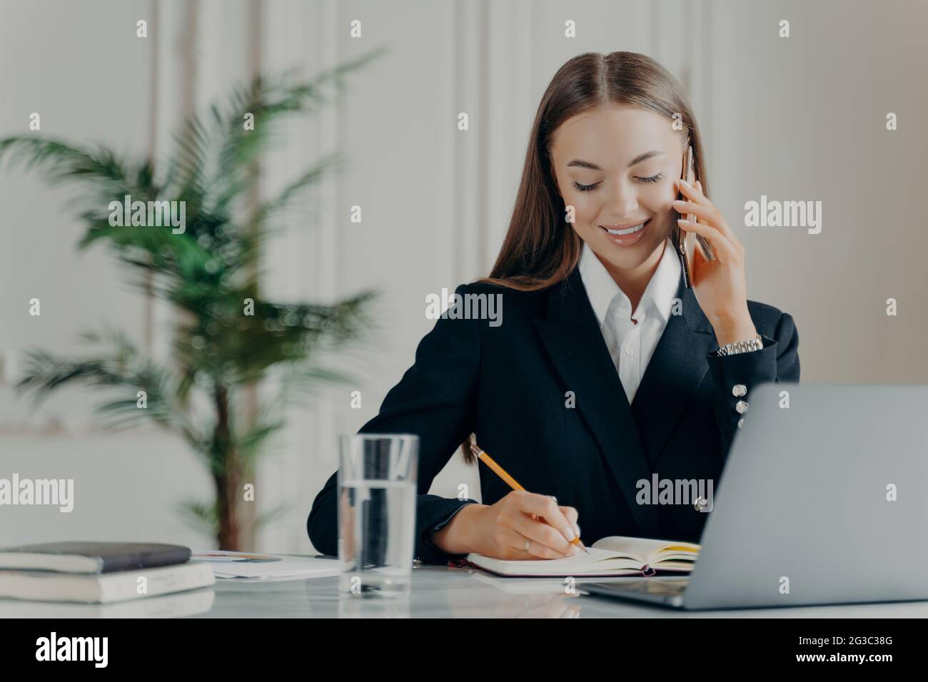 Positive junge Geschäftsfrau, die telefoniert und Notizen schreibt Stockfoto