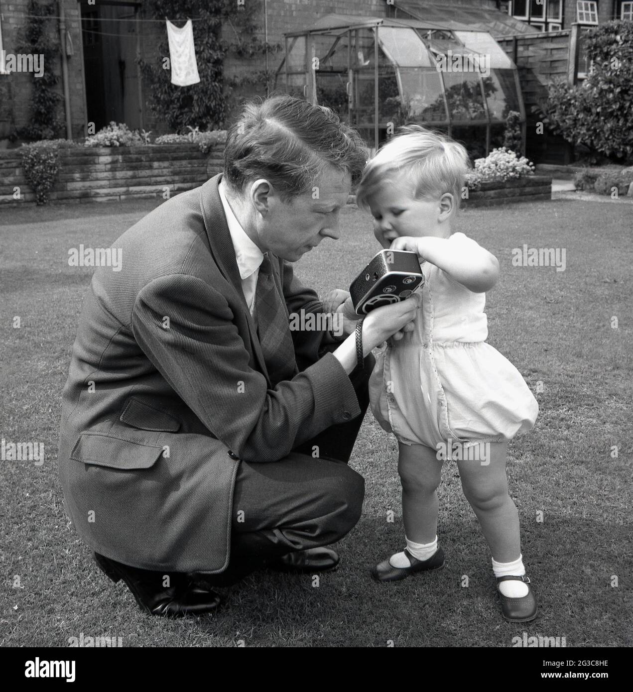 In den 1950er Jahren, historisch, draußen in einem Garten, kniete ein Vater in Anzug und Krawatte nieder und zeigte seinen kleinen Sohn, seine kleine, handgehaltene 8-mm-Cine-Kamera, England, Großbritannien. Solche leichten Cine-Kameras mit 8mm-Film waren in dieser Ära ein Popula-Hobbyr, der verwendet wurde, um sogenannte „Heimfilme“ zu drehen, die Aufzeichnungen über das gewöhnliche Leben der Menschen drehten. Stockfoto