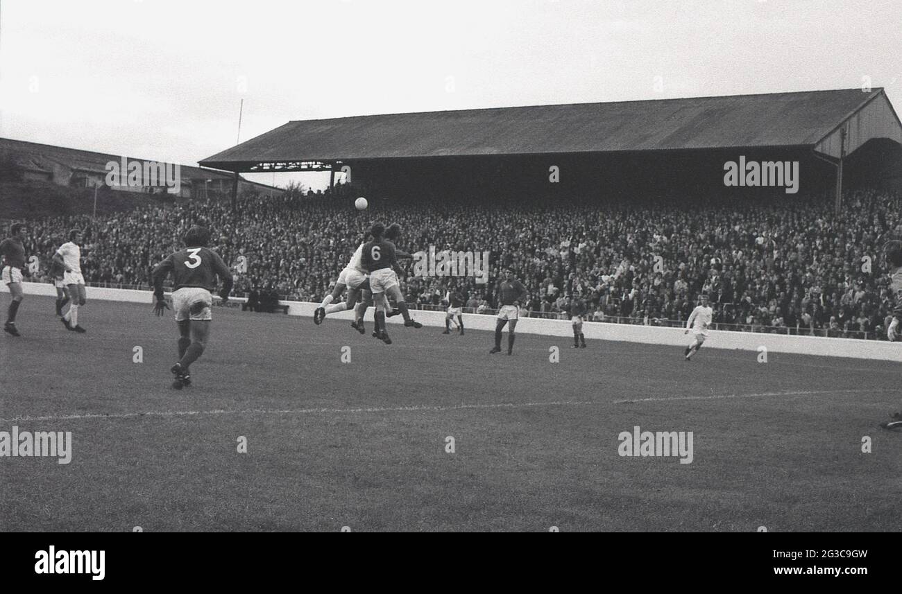 1970s, historisch, ein Fußballspiel, Millwall verus Charlton, auf ihrem Boden, The Den, Cold Blow Lane, Surrey Docks, East London, England, Großbritannien, wo der FC Millwall von 1910 bis 1993 spielte. Das Bild zeigt die Spieler, die um den Ball kämpfen, und die halbwegs überdachte Terrasse mit Teiltribünenabdeckung, die Nordterrasse, die voller Zuschauer ist. Das von Archibald Leitch entworfene „The Den“ war 83 Jahre lang die Heimat des FC Millwall und ein feindseliger, einschüchternder Austragungsort gegnerischer Teams. Durch die Luftangriffe von WW2 schwer beschädigt, wurde es mit Hilfe von Unterstützern wieder aufgebaut und 1944 wiedereröffnet. Stockfoto