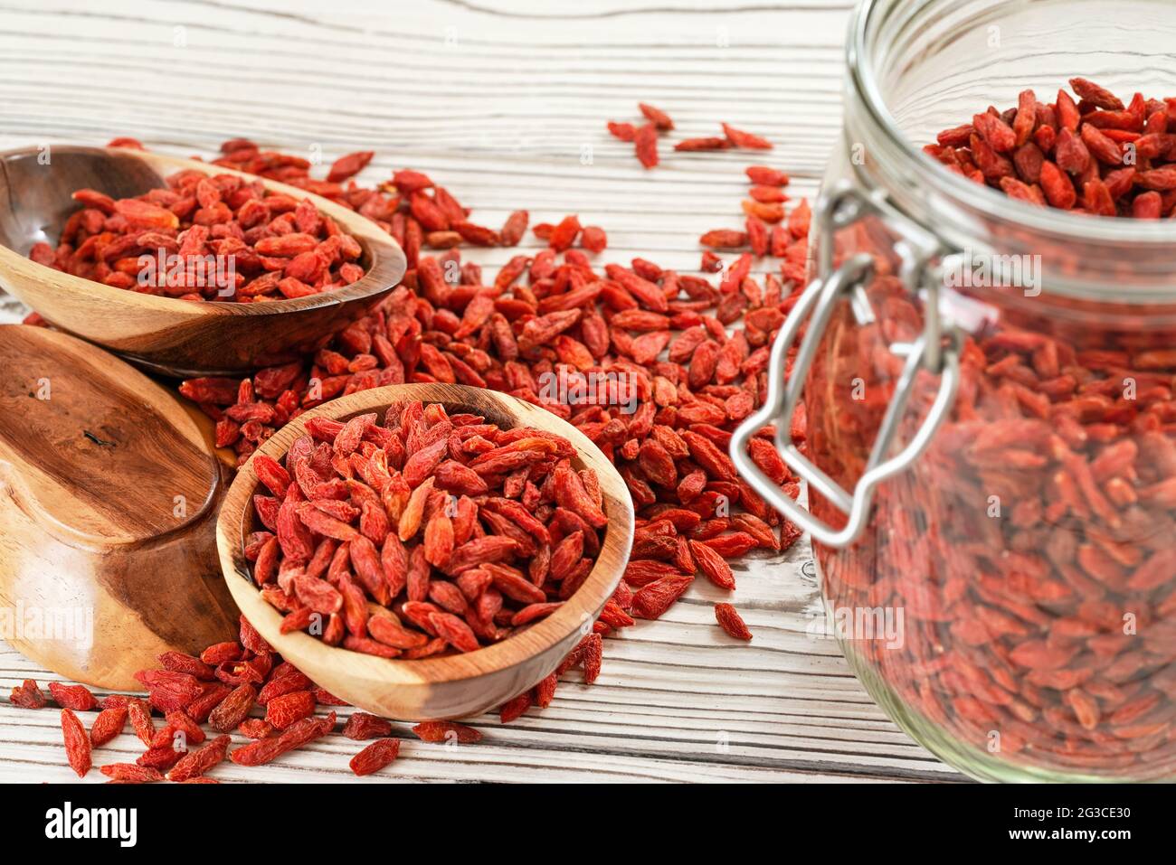 Getrocknete Goji aka. wolfberry Samen in Glasflasche, einige in Holzschüsseln und verschüttet auf weißen Brettern Schreibtisch Stockfoto