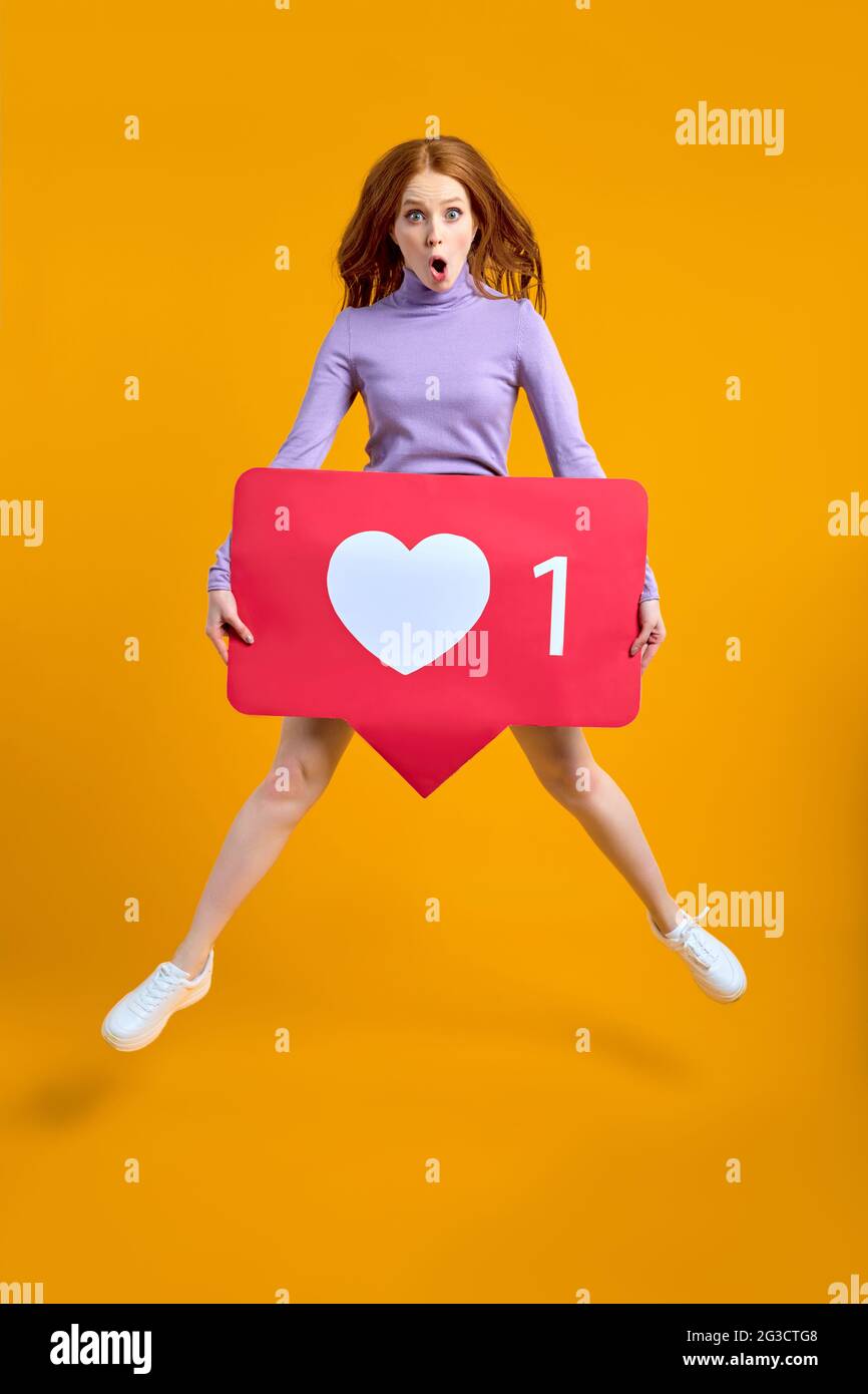 Schockiert springende Frau mit Herzknopf, rothaarige Frau hält wie Symbol der sozialen Medien, Emoji-Zeichen zu folgen abonnieren interessante Blog, beliebt fo Stockfoto