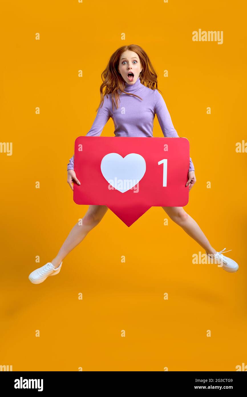 Überrascht springende Frau mit Herzknopf, Rotschopf weiblich halten wie Symbol der sozialen Medien, Emoji-Zeichen zu folgen abonnieren interessante Blog, beliebt Stockfoto