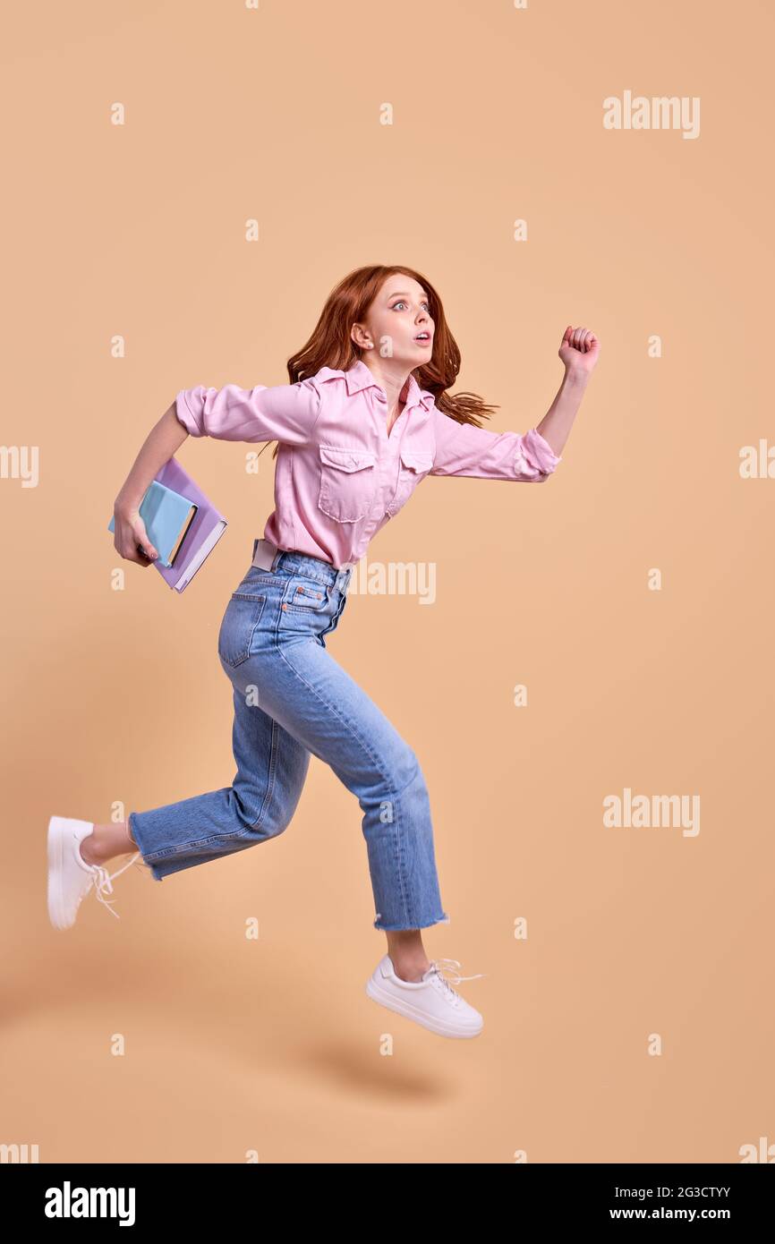 Lustige Rotschopf-Studentin eilt mit Buch in den Händen vorwärts, eilt zur Universität, trägt lässiges Outfit, läuft, Prüfung am 1. September, Isoliert in s Stockfoto