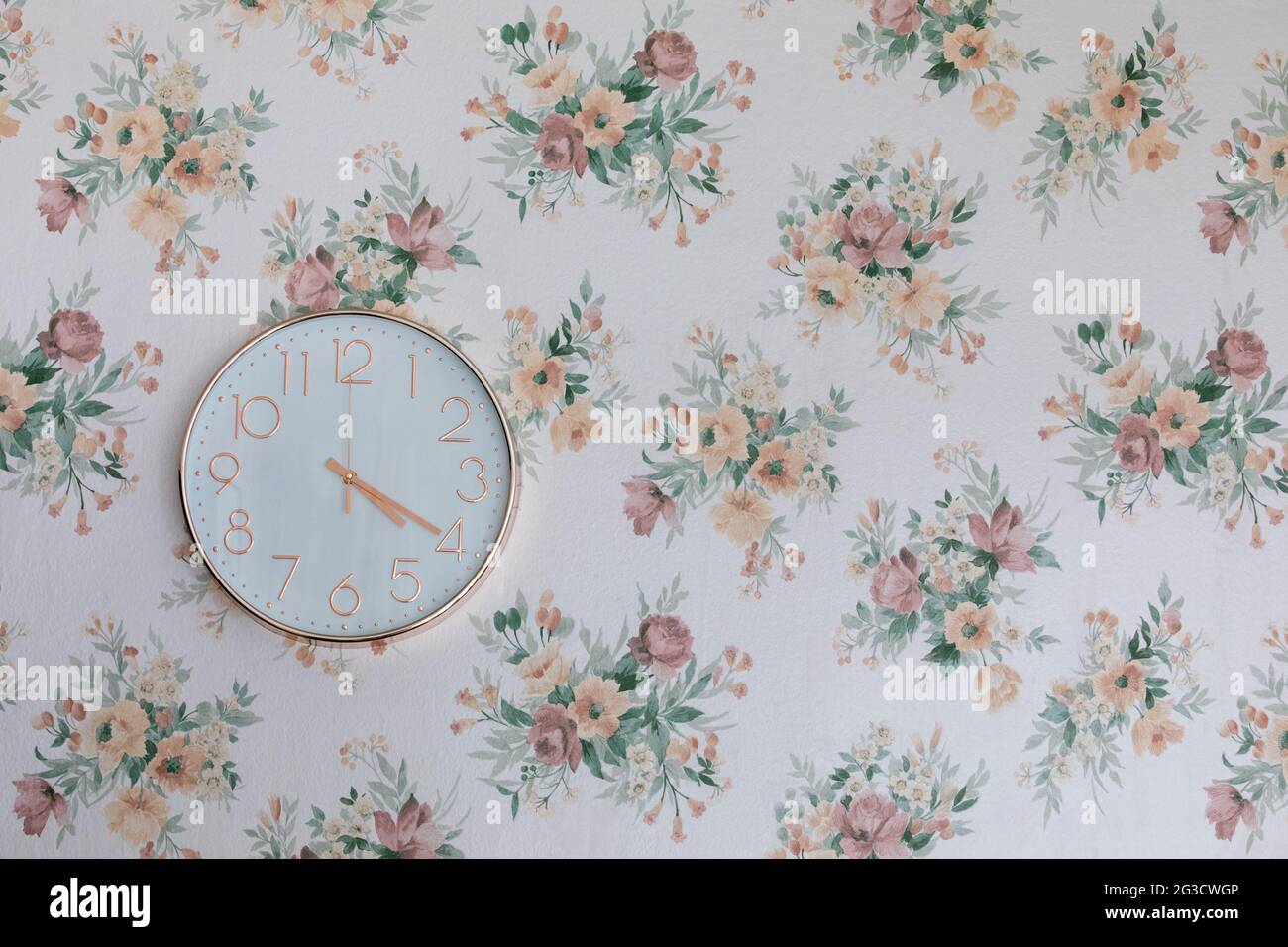 Roségoldfarbene Uhr in runder Form, an einer Wand mit floraler Tapete montiert Stockfoto