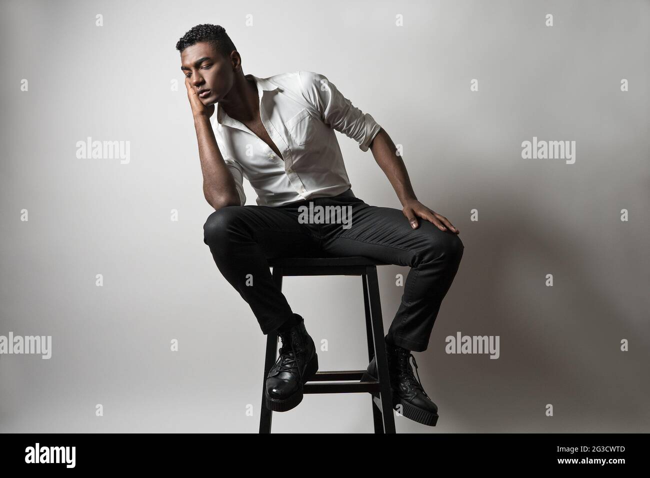 Ein junger schwarzer Mann, Model, sitzt auf einem Holzhocker, lehnt mit geschlossenen Augen, entspannt und denkend. Ein künstlerisches Porträt. Stockfoto