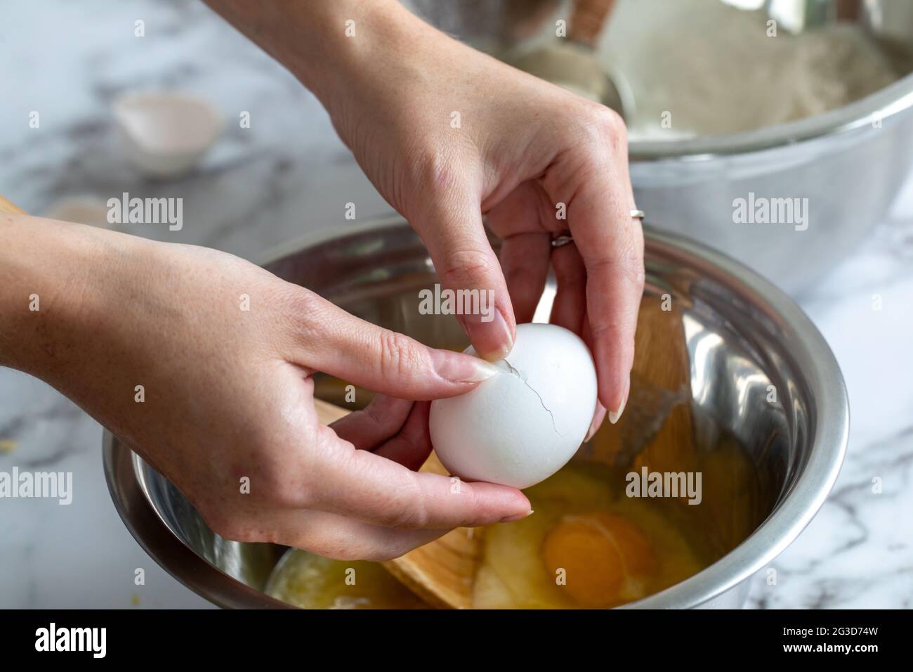 Die Hand einer Frau, die ein Ei über einer gewickelten Metallmischschüssel mit natürlichem Licht aufbricht Stockfoto