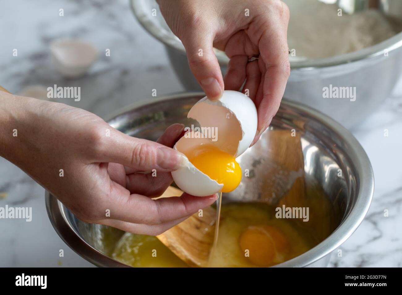 Die Hand einer Frau, die mit dem gelben Eigelb ein Ei aufbricht, über einer runden Metallschüssel mit natürlichem Licht Stockfoto