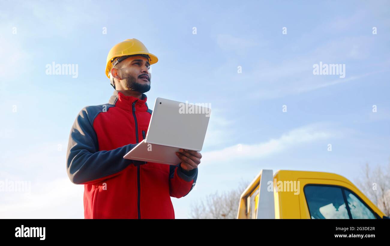 Junger Arbeiter mit einem gelben Schutzhelm, der einen Laptop in der Hand hält und nach einer Adresse sucht. Hinter ihm ist ein gelber LKW zu sehen. Blauer Himmel im Hintergrund. Tagesaufnahme in hoher Qualität. Stockfoto