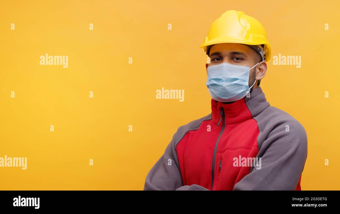 Porträt eines jungen indischen Ingenieurs mit einem gelben Schutzhelm und einer Maske, die im Studio mit gekreuzten Armen auf gelbem Hintergrund steht. Isolierter Mann. Nahaufnahme. Stockfoto