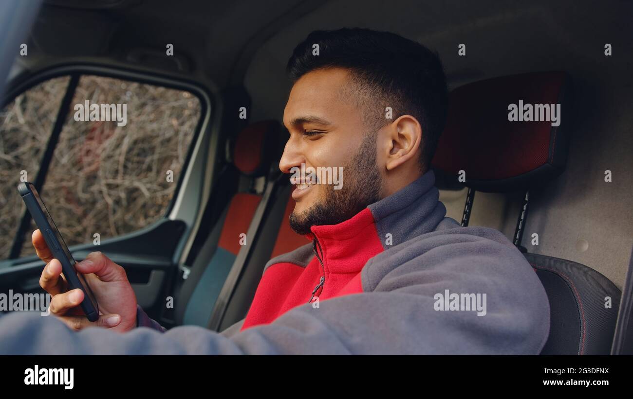 Junger Lieferer sitzt auf dem Vordersitz des Wagens mit der Hand am Steuer. Hält ein Telefon in der anderen Hand und lächelt. Über sein Telefon. Trägt eine rote Uniform. Kurierkonzept. Stockfoto