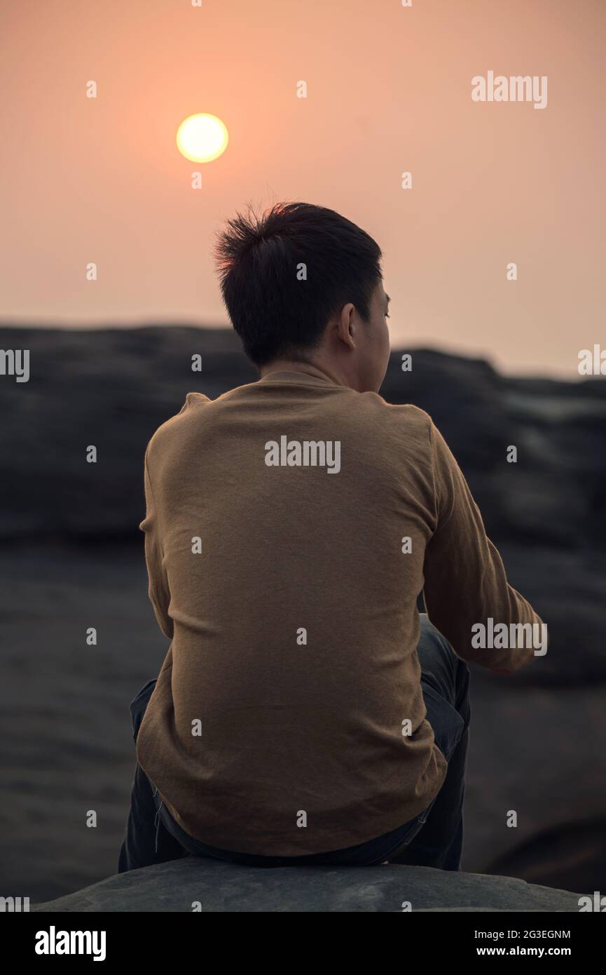 Einsamkeit junger asiatischer Mann, der beim Sonnenuntergang etwas auf einer Felsklippe ansieht Stockfoto