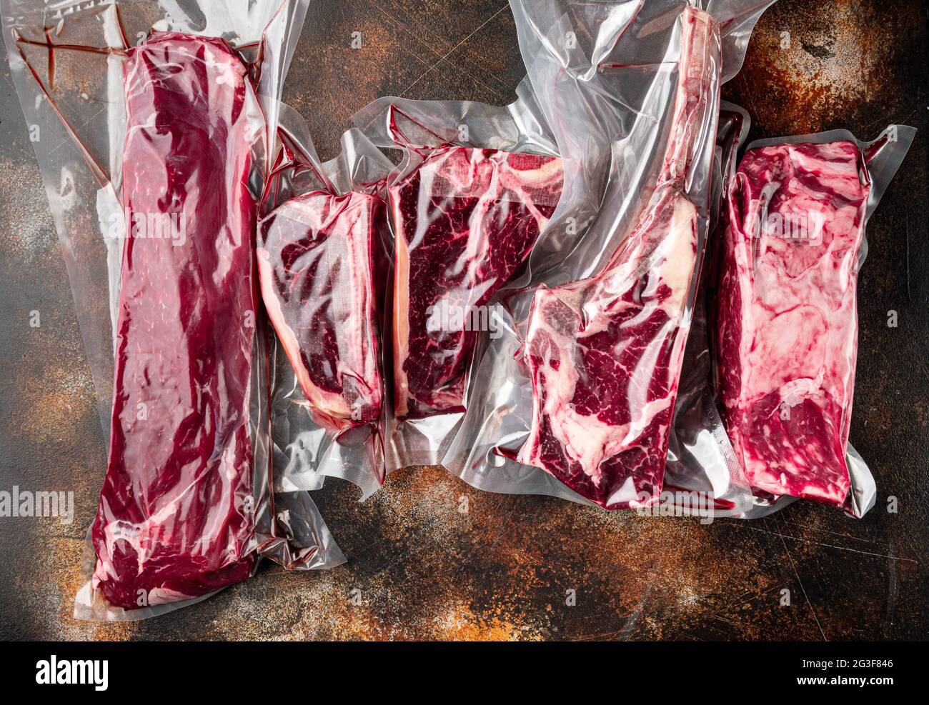 Rohes marmoriertes Rindersteak im Vakuumverpackungsset, Tomahawk, T-Bone, Clubsteak, Rib-Eye- und Filet-Schnitte, auf altem dunklen rustikalen Hintergrund, Draufsicht Fla Stockfoto