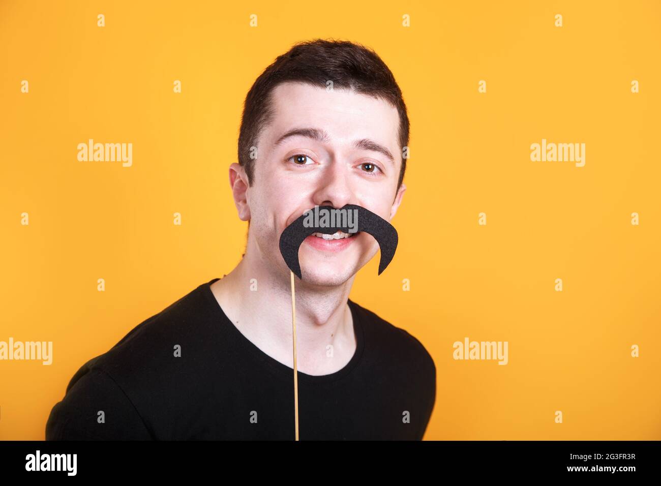 Lustiger junger Mann, der einen gefälschten Schnurrbart in einem Stock vor seinem Gesicht auf gelbem Hintergrund hält Stockfoto