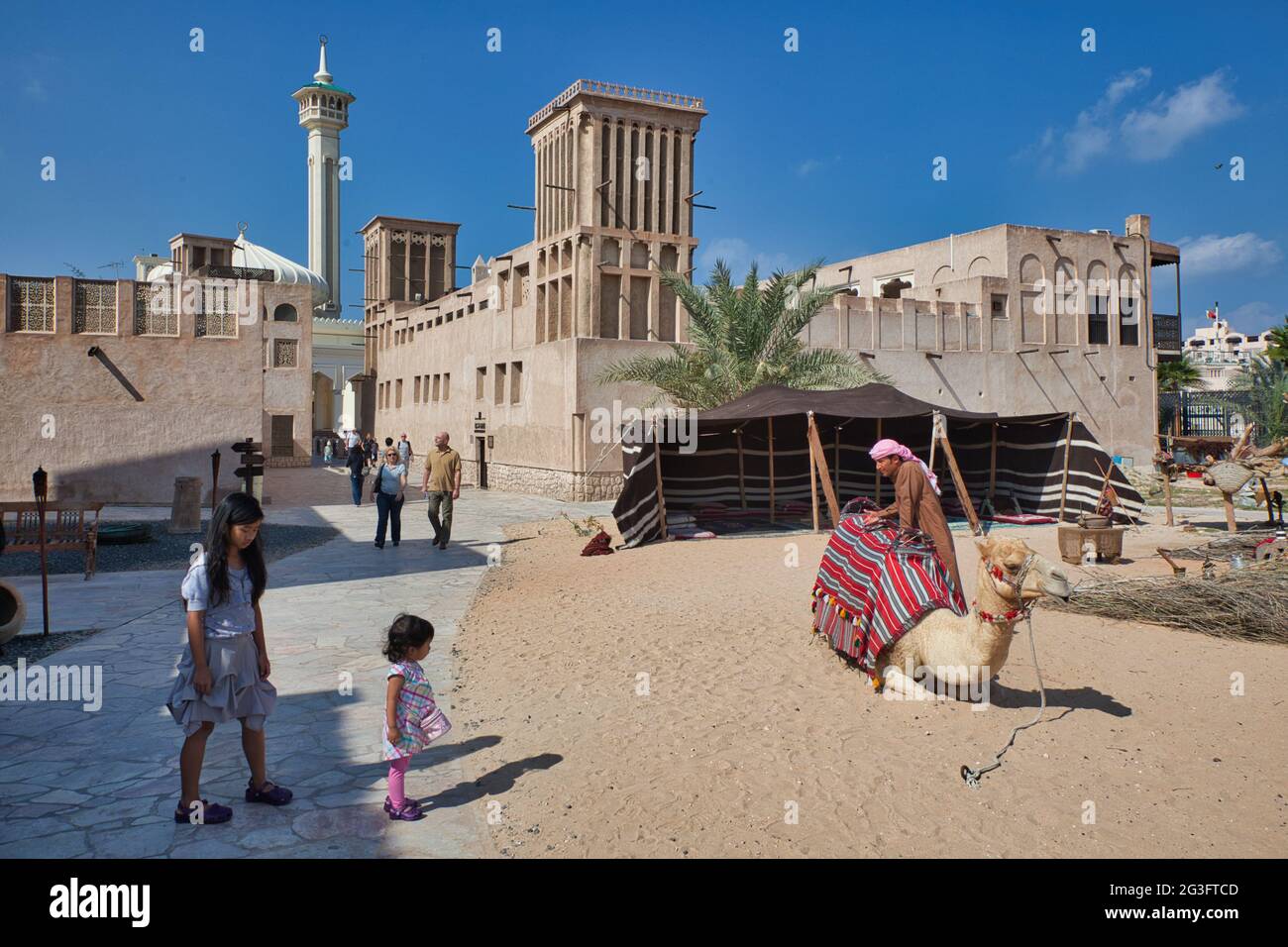Eine Mutter und eine kleine Tochter, die einen Kamel- und Kamelbesitzer in der Altstadt von Dubai, den Vereinigten Arabischen Emiraten, mit einem Haus mit einem kühlenden Windturm gleich dahinter anschaut Stockfoto