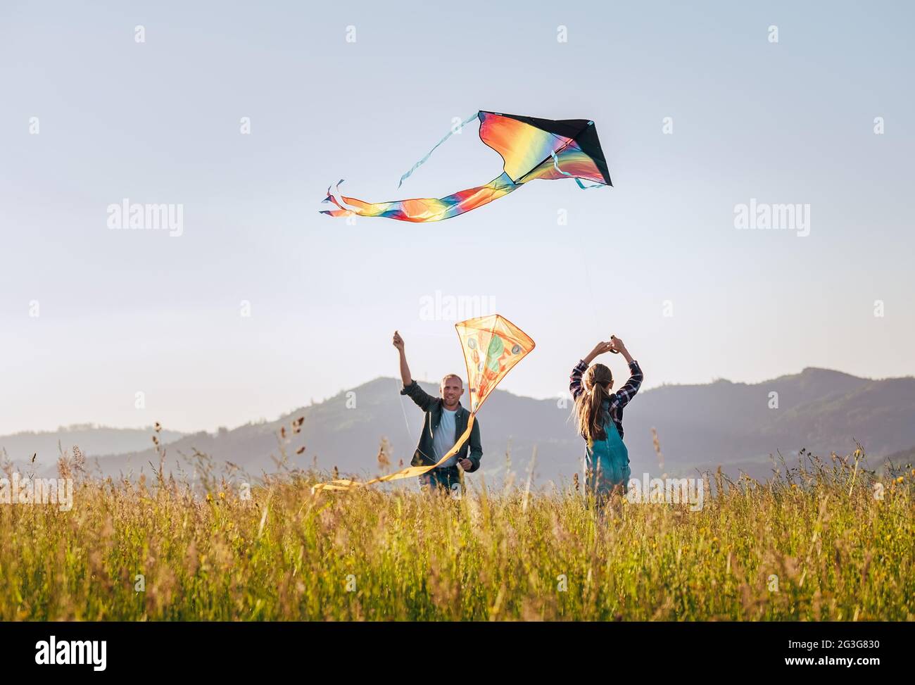 Tochter mit lächelndem Vater, während sie eine bunte Drachen auf der hohen Graswiese in den Bergfeldern fliegen. Warme Momente für die Familie oder im Freien Stockfoto