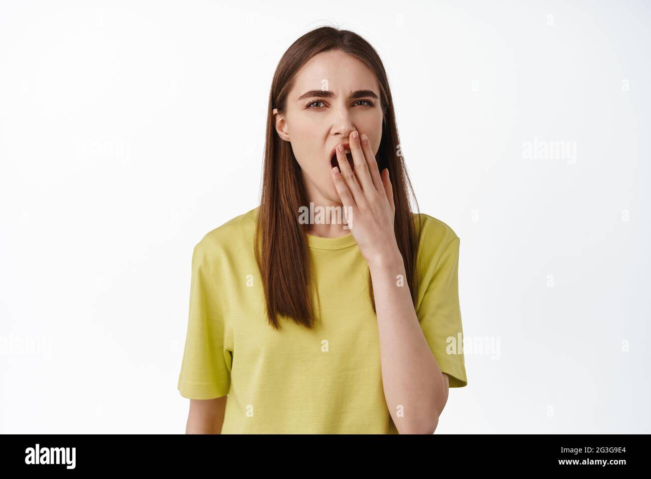 Porträt einer müden jungen Frau, die gähnt, den geöffneten Mund mit der Hand bedeckt und gähnt, sich erschöpft fühlt, sich vom Smth gelangweilt, im gelben T-Shirt dagegen stehend Stockfoto