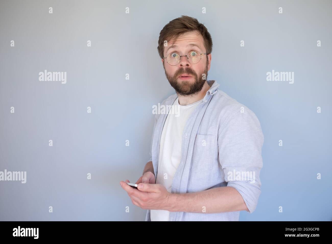 Verwirrt, schockiert Mann mit einem Smartphone in den Händen schaut auf die Kamera überrascht, mit großen Augen. Grauer Hintergrund. Stockfoto