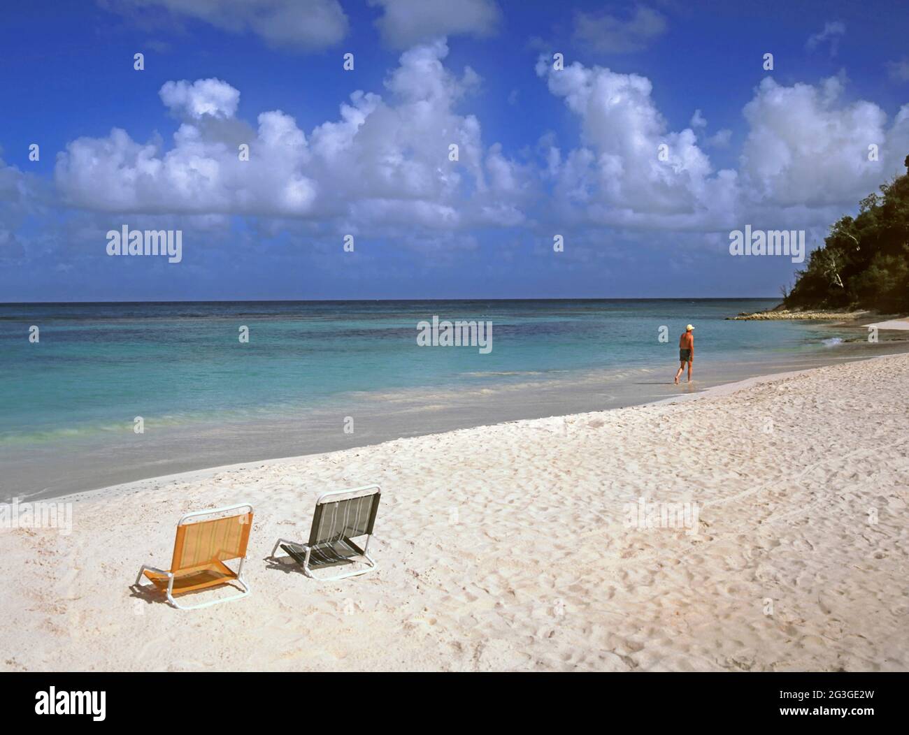Rückansicht des Lebensstils eines Mannes allein, der in klarem, ruhigen türkisfarbenen Wasser der Karibik paddelt und spazierengeht Die Küste hinterlässt zwei Stühle an einem Sandstrand mit weißen Cumulus-Wolken am blauen Himmel über dem Meereshorizont Die Küste von Long Bay Beach Antigua West Indies Leeward Islands Karibik Stockfoto