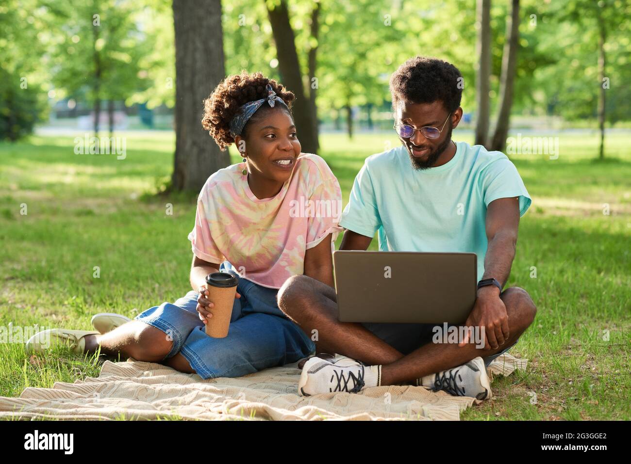 Afrikanisches junges Paar, das auf dem Gras sitzt, Kaffee trinkt und im Park einen Laptop benutzt Stockfoto