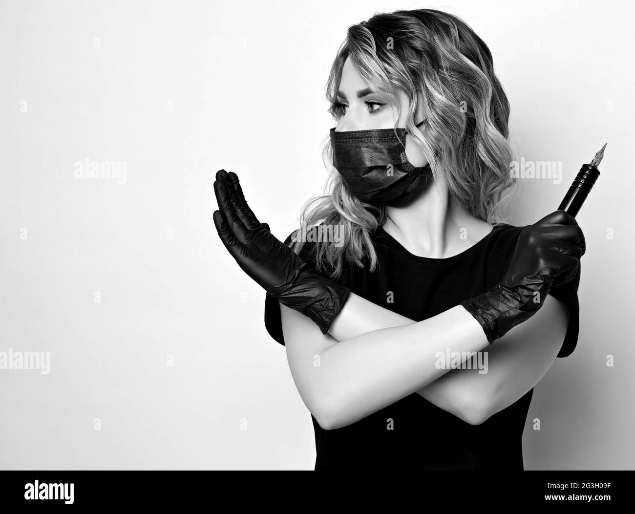 Schwarz und Weiß. Lockige Frau in schützende medizinische Maske und Handschuhe mit Permanent Make-up-Maschine in Hand Gesten Kreuz Stockfoto