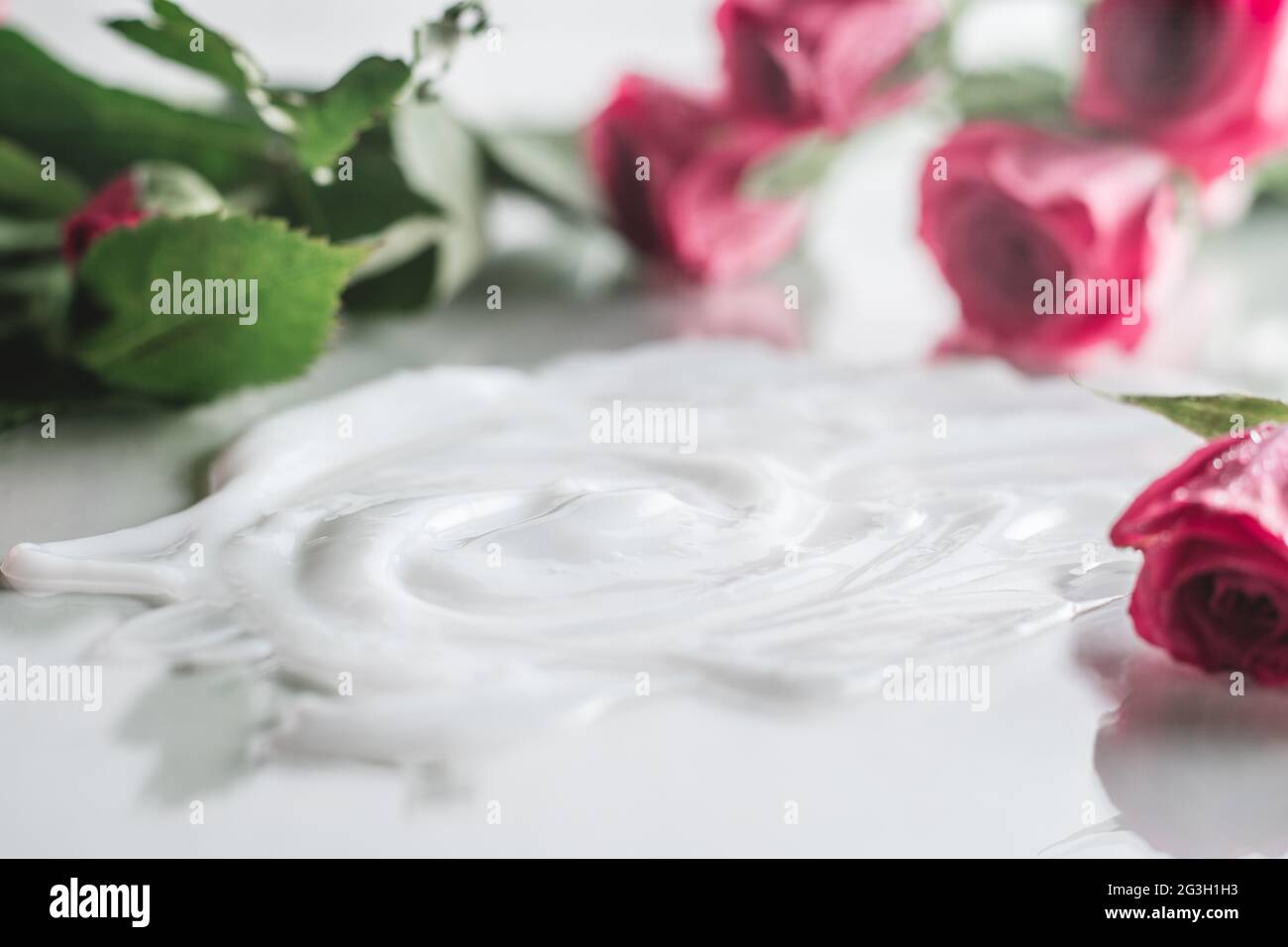 Weiße Creme auf glänzender Oberfläche, umgeben von unscharfen, zarten rosa Rosen Stockfoto