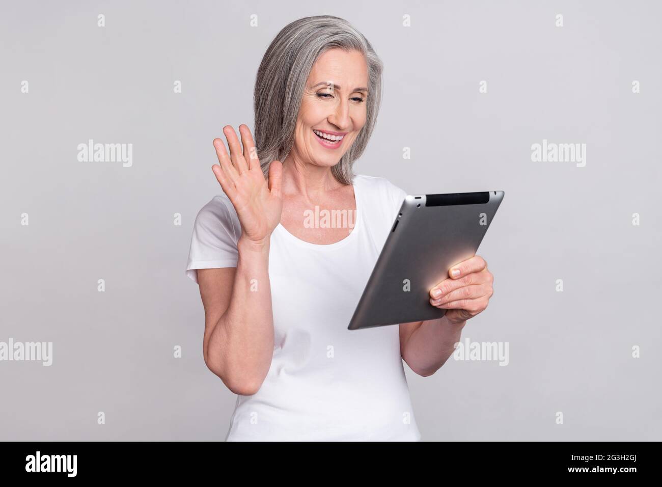 Foto von älteren Frau glücklich positive Lächeln sprechen Welle Hand hallo Videoanruf Tablet isoliert über grauen Hintergrund Stockfoto