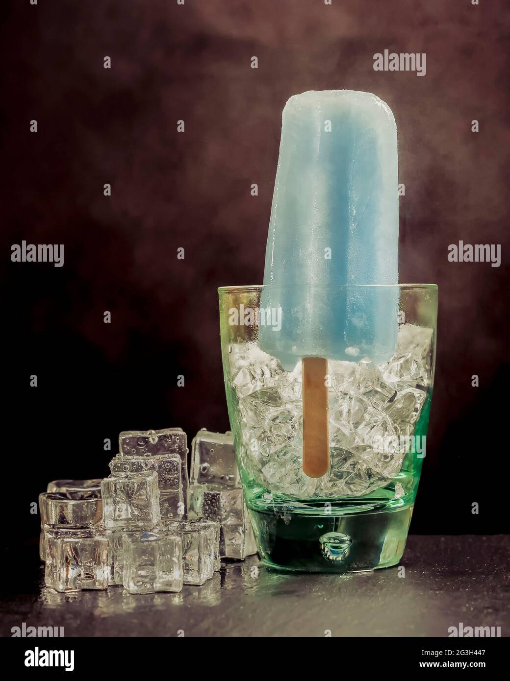 Ein blauer Popsicle steht in einem Glas mit Eis und Rauch auf dem dunklen Hintergrund Stockfoto