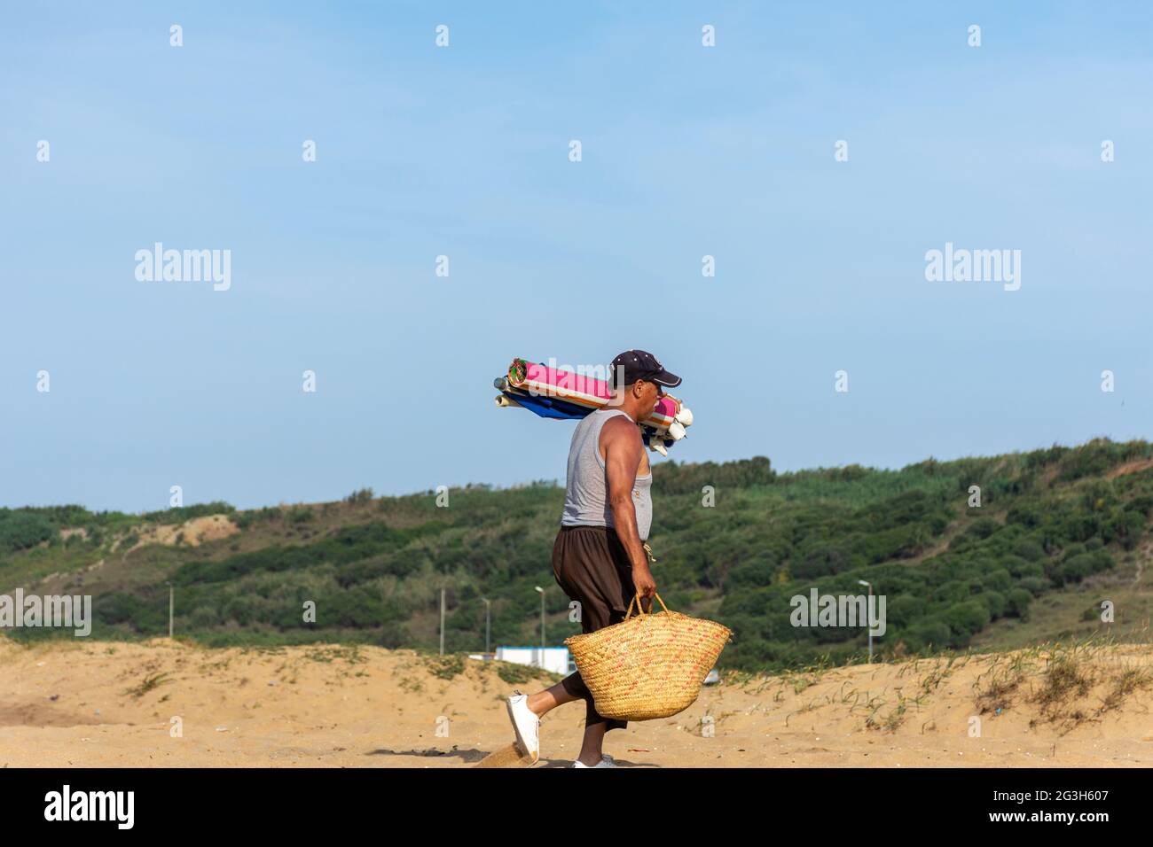 Ein Algerier mittleren Alters, der einen traditionellen französischen Marktkorb und Teppiche am Strand hält. Stockfoto