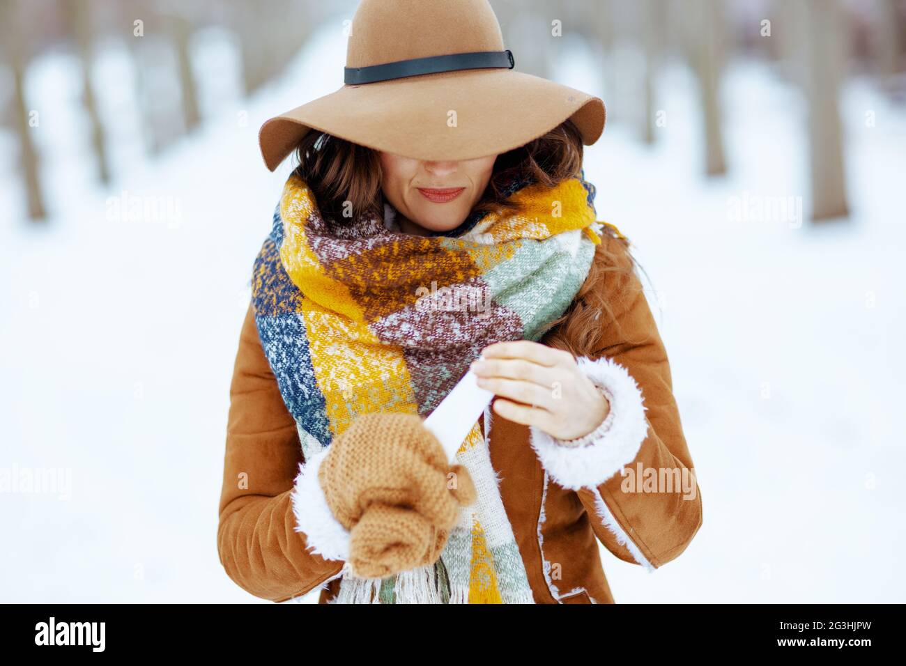 Lächelnde elegante Frau in braunem Hut und Schal mit Fäustlingen in  Schaffell Mantel mit Gesichtscreme im Freien im Stadtpark im Winter  Stockfotografie - Alamy