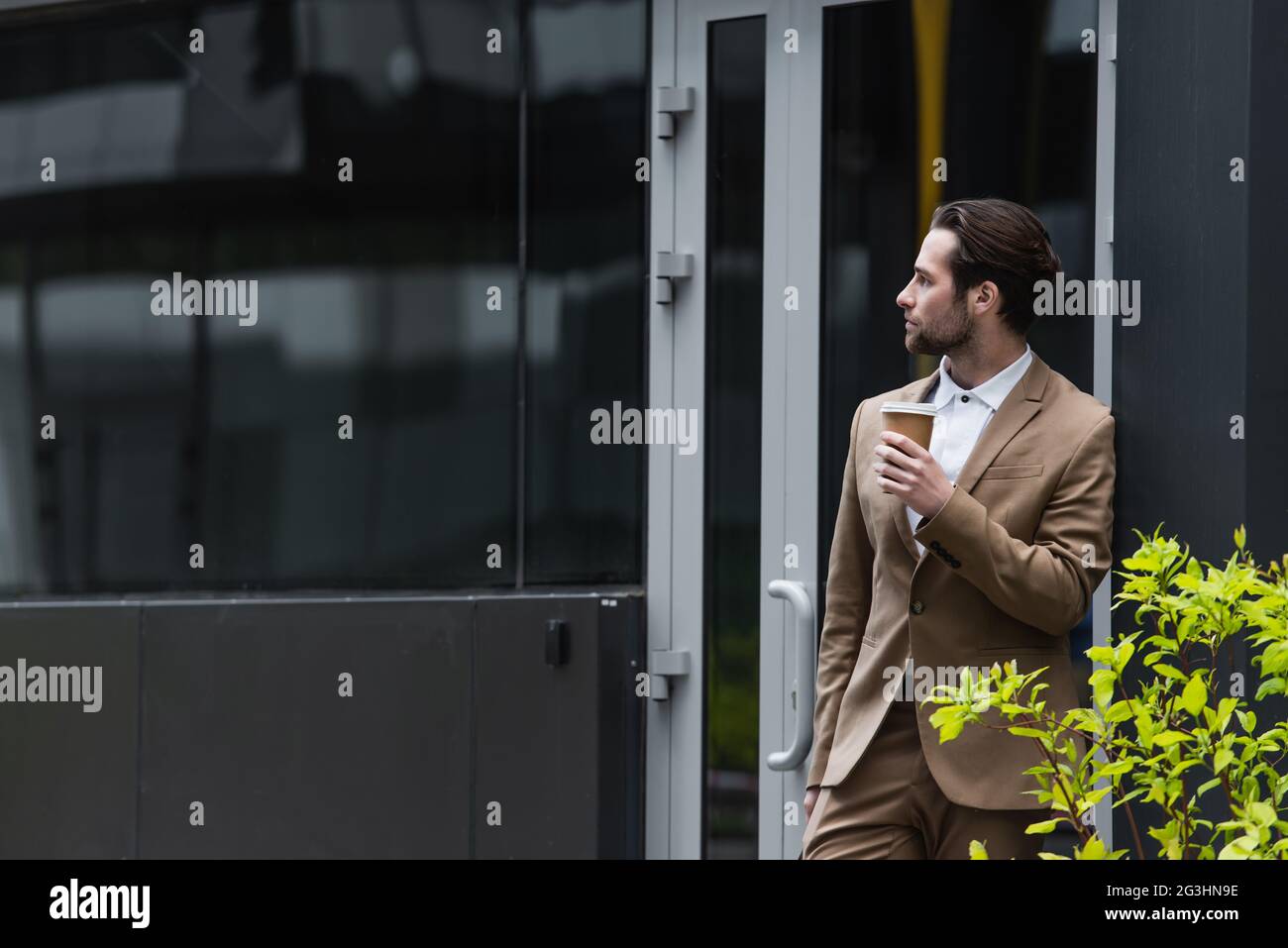 Nachdenklicher Geschäftsmann in formeller Kleidung, der Kaffee hält, um in die Nähe des Gebäudes zu gehen Stockfoto