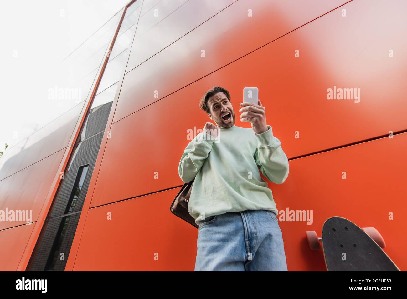 Low-Angle-Ansicht des aufgeregten Mannes in einem Sweatshirt, das das Smartphone in der Nähe von Longboard und orangefarbener Wand hält Stockfoto
