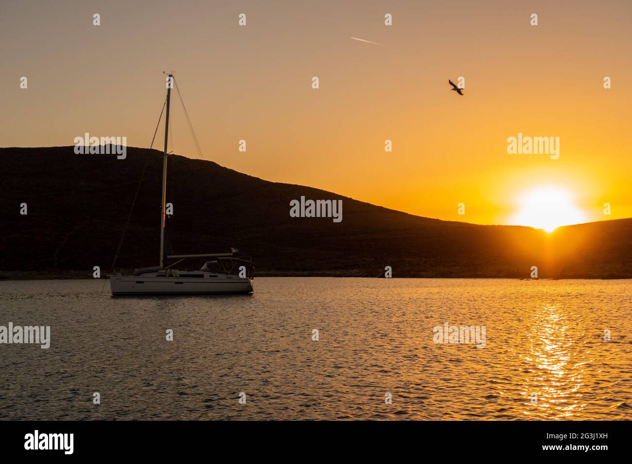 Magischer, lebhafter orangefarbener Sonnenuntergang über dem Hügel und dem Mittelmeer mit einer Silhouette eines Segelbootes, während der Vögel fliegen, in einer Bucht auf der Insel Rineja Stockfoto