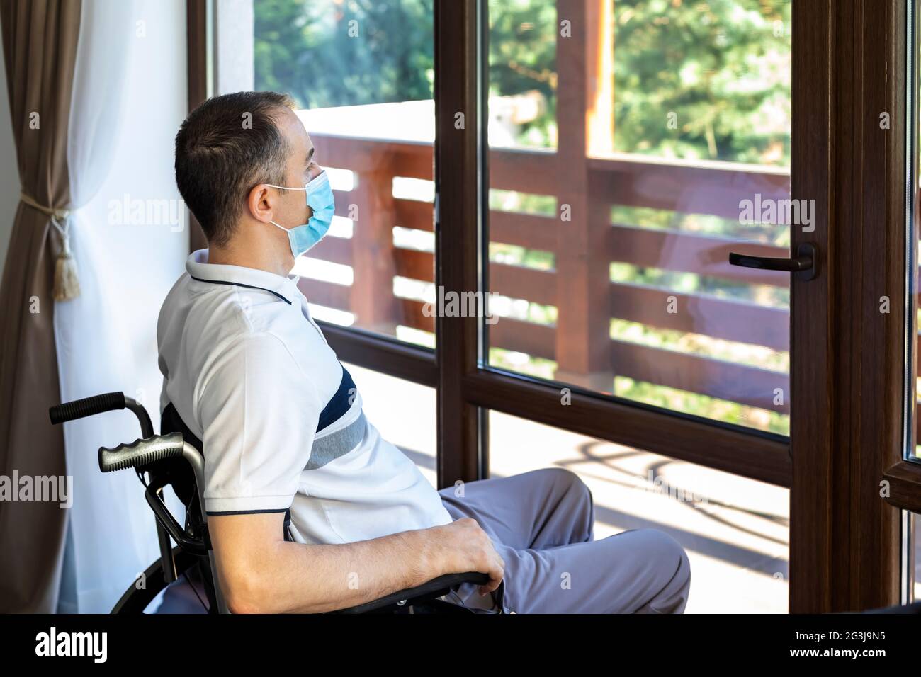 Einsamer junger Mann mit Gesichtsmaske sitzt allein in einem Rollstuhl und schaut aus dem Fenster. Konzentrieren Sie sich auf sein Gesicht. Stockfoto