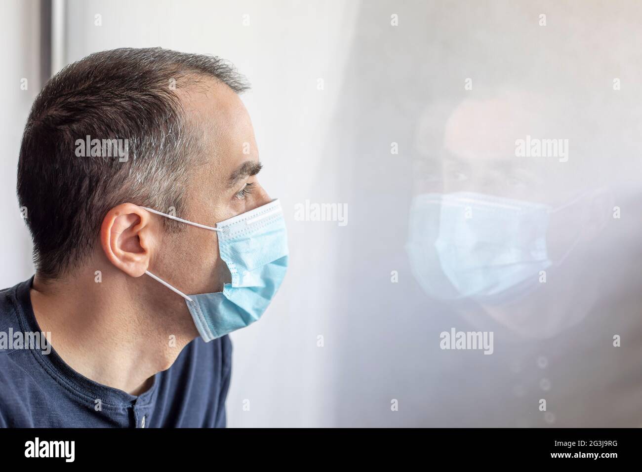 Junger Mann mit medizinischer Schutzmaske und Blick aus dem Fenster. Mann in Isolation zu Hause. Stockfoto