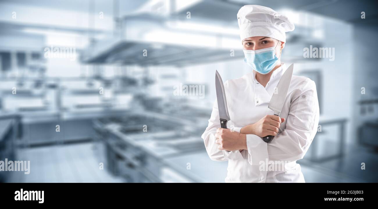 Panorama-Banner eines Küchenchefs mit Maske aufgrund einer covid-19 Pandemie, der mit gekreuzten Händen stand und zwei scharfe Messer vor dem Restaurantkit hielt Stockfoto