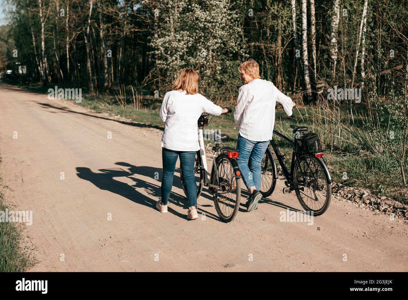 Zwei reife Freundinnen laufen mit Fahrrädern entlang des Waldes. Rückansicht Stockfoto