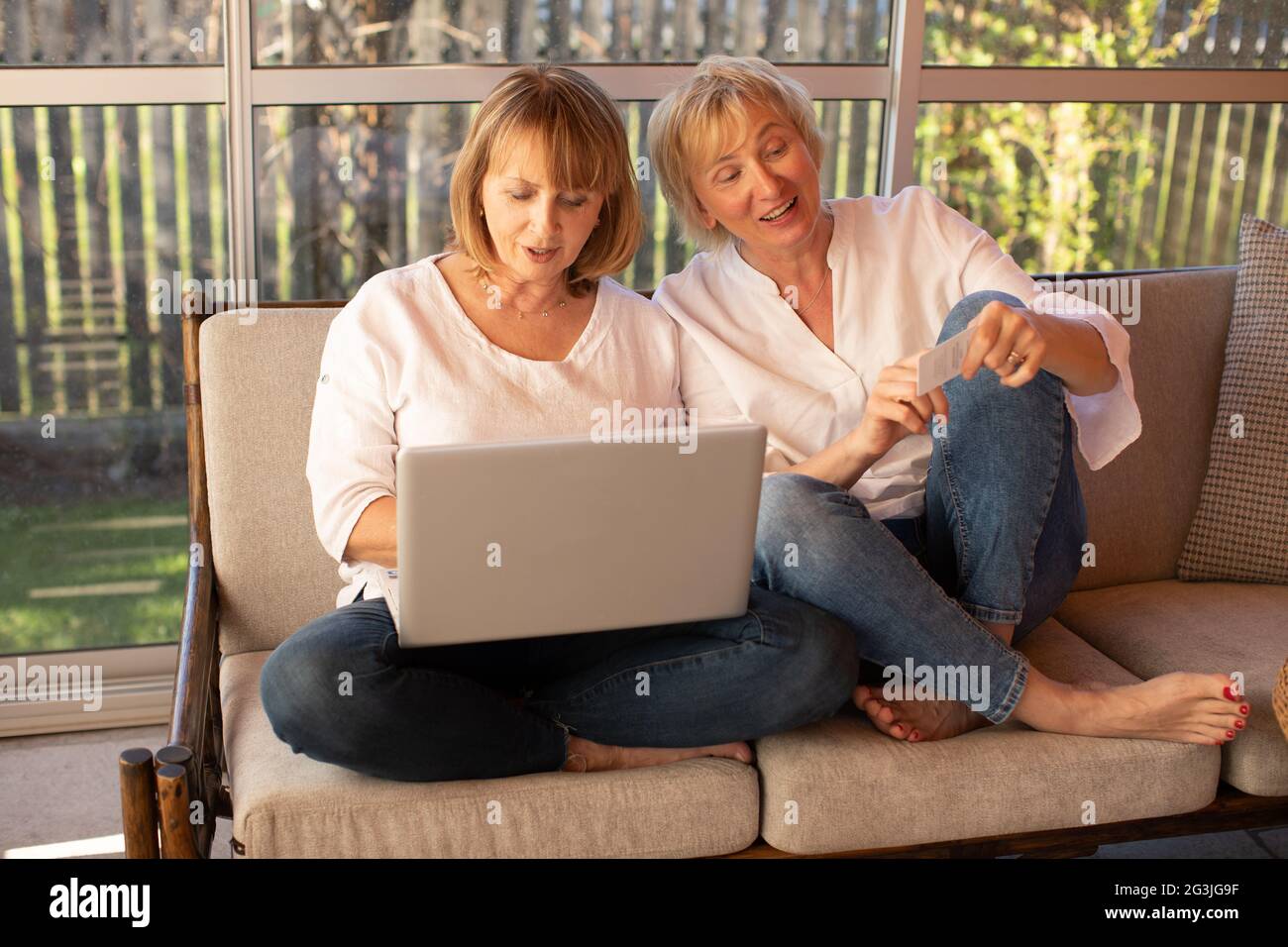 Zwei 55-jährige Frauen in legerer Kleidung kaufen mit einem Laptop online, während sie bequem auf der Couch zu Hause sitzen Stockfoto
