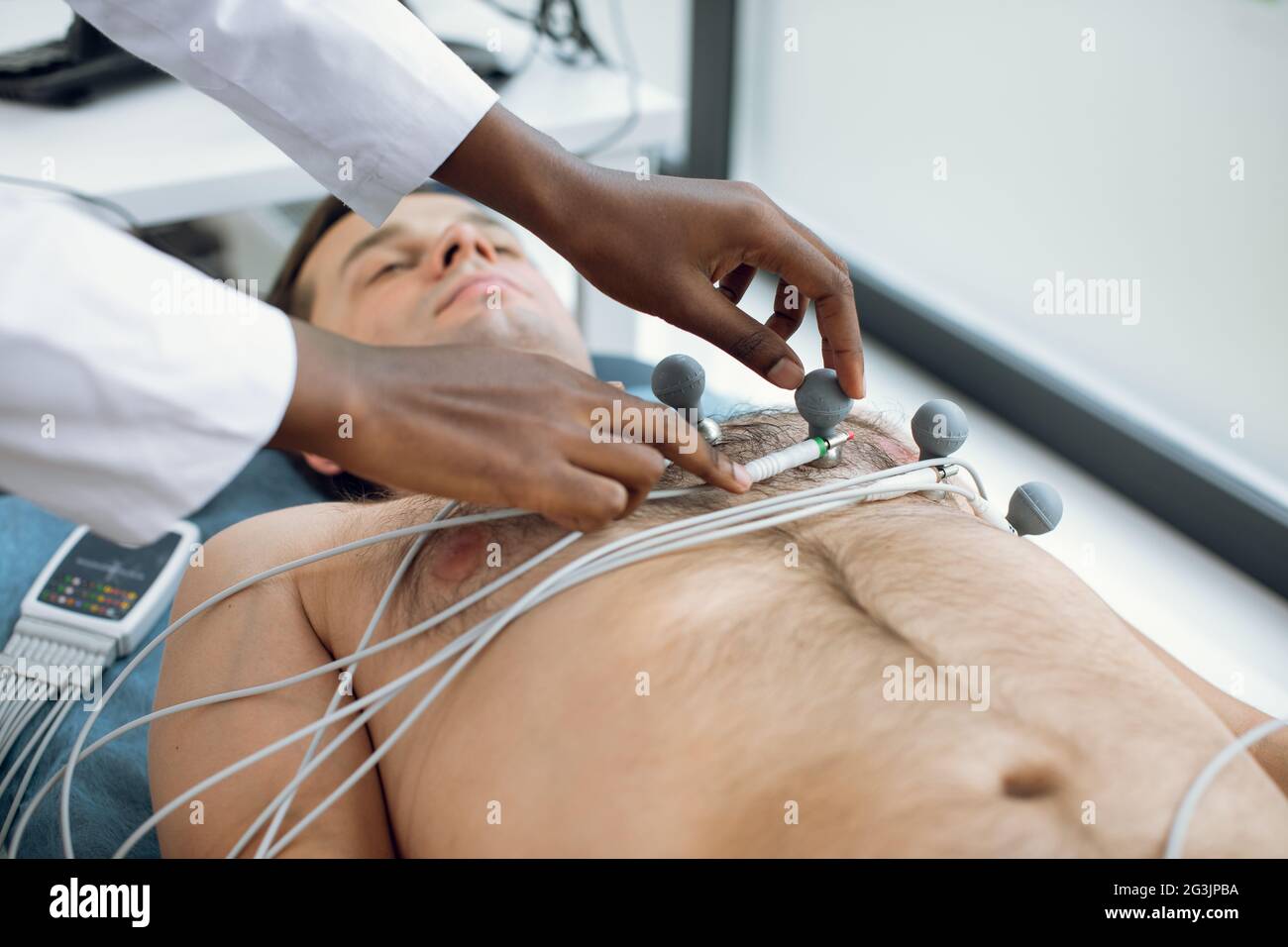 Zugeschnittenes Bild einer afroamerikanischen Ärztin oder Krankenschwester, die im Krankenhaus holter- und Vakuumelektroden auf die Brust eines jungen männlichen Patienten klebte. EKG- und Herzgesundheitskonzept. Jährliche Integritätsprüfung Stockfoto