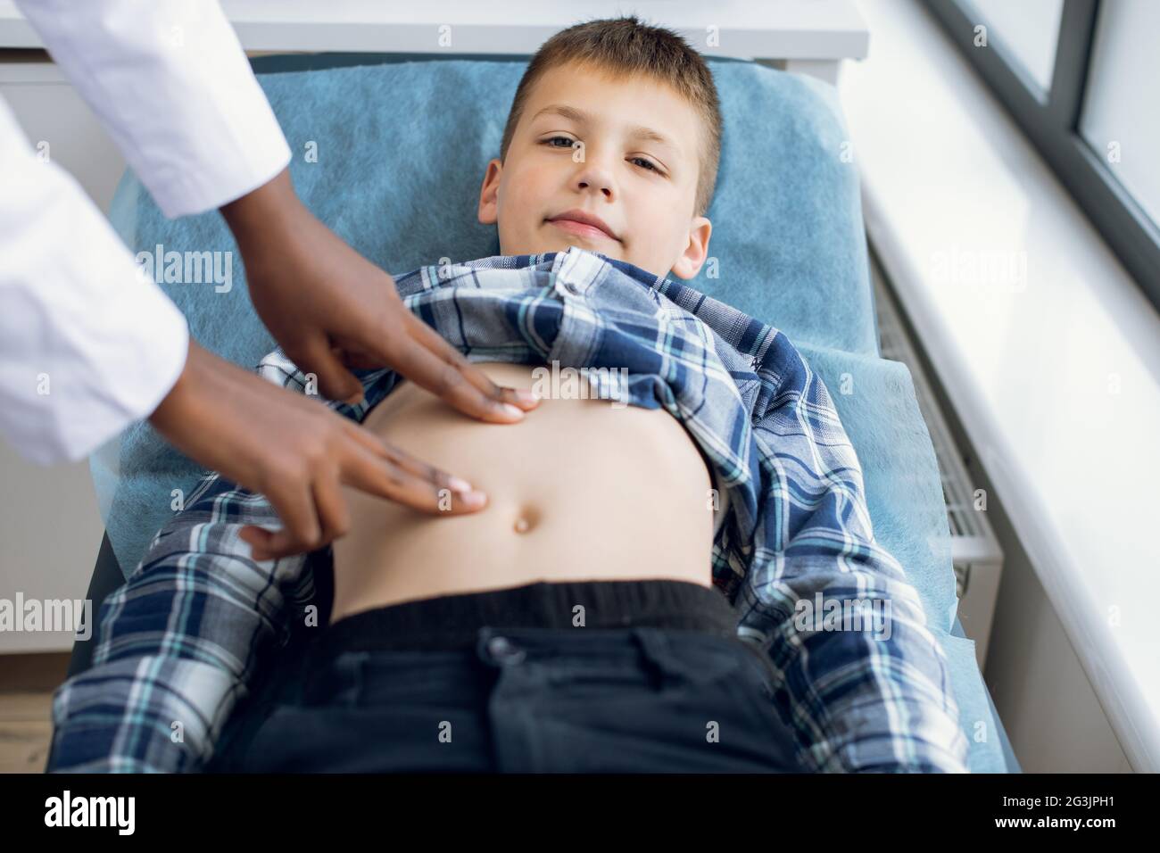 Netter lächelnder Junge im Alter von 10 Jahren, der auf der Couch in der modernen Klinik liegt und seinen Arzt besucht. Weibliche Afro-Kinderärztin, die den Abdomen eines Kindes mit manueller Palpation untersucht Stockfoto