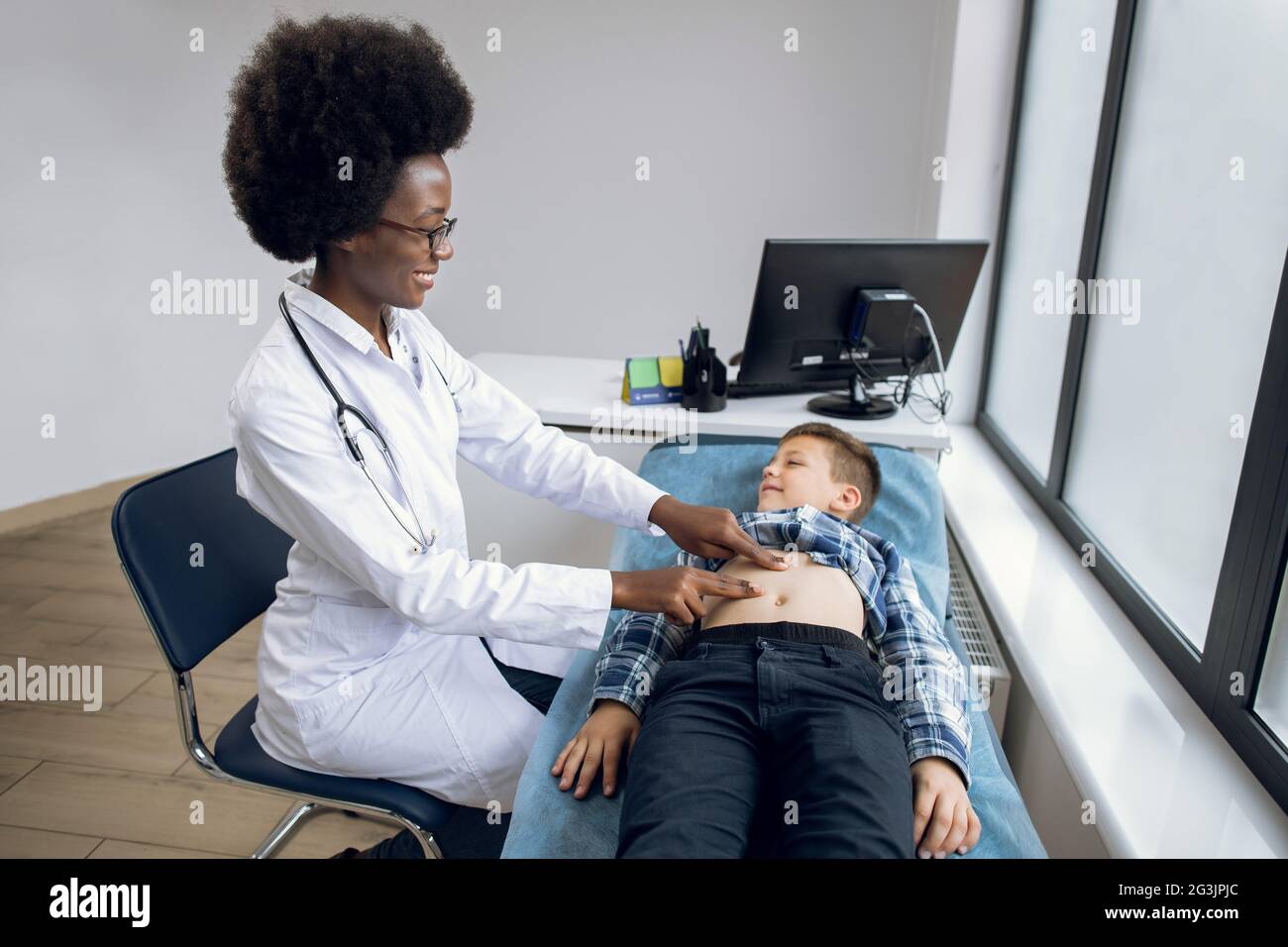 Porträt einer lächelnden, fröhlichen afrikanischen Ärztin, die manuelle Palpation durchführt, den Bauch eines kleinen Jungen untersucht und mit ihm spricht, wo es weh tut. Bauchschmerzen Konzept Stockfoto