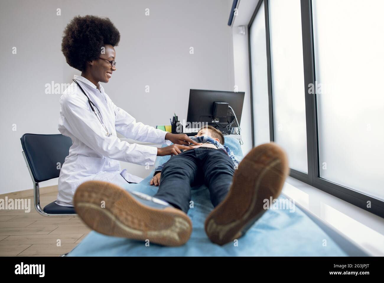 Afroamerikanische Ärztin gp Kinderärztin, im weißen Mantel, der den Bauch eines jungen Teenagers klopfend, auf der Couch liegend, lächelnd und mit einem kleinen Patienten sprechend. Bauchschmerzen, medizinisches jährliches Kontrollkonzept Stockfoto
