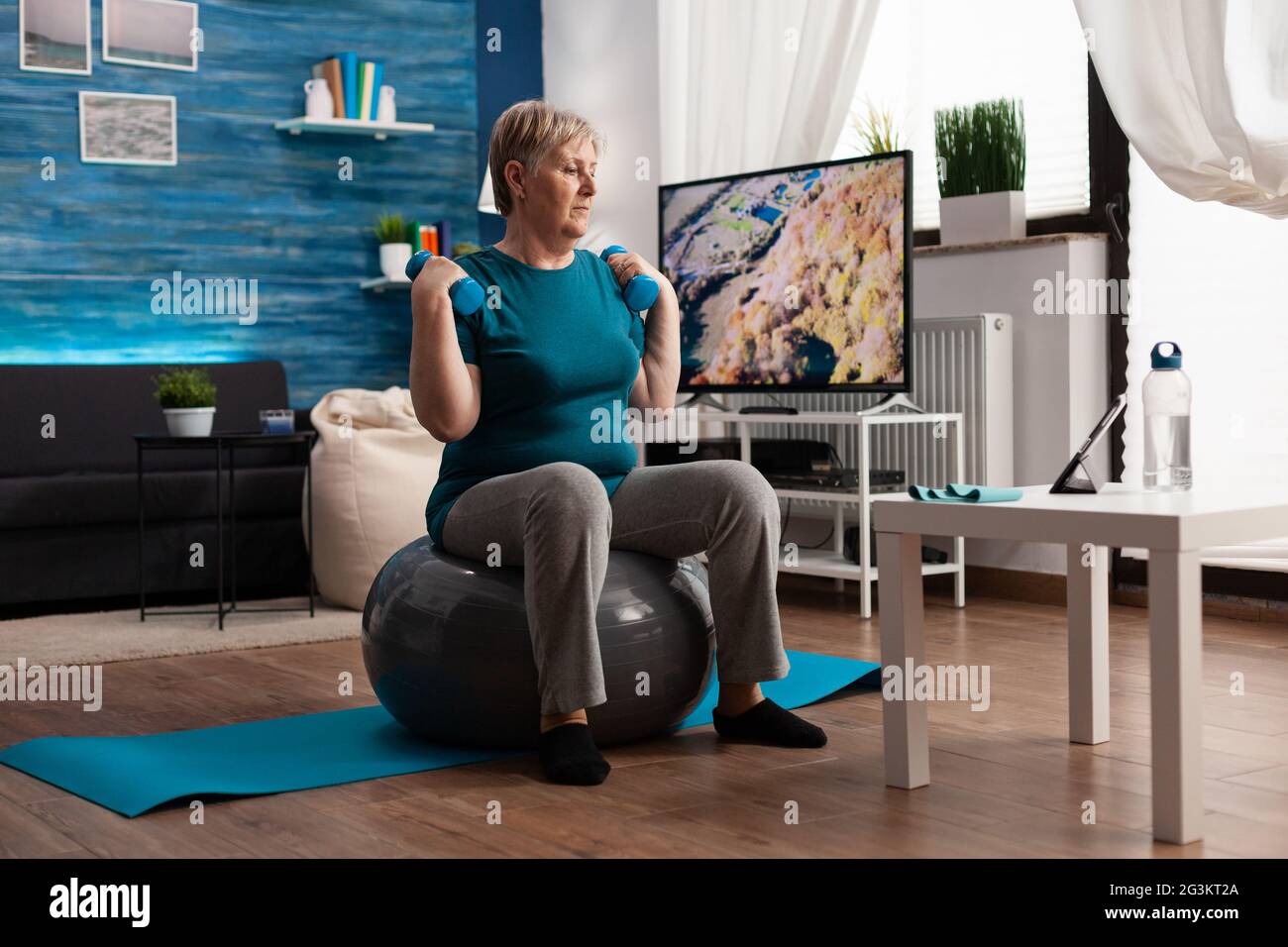 Fokussierter Senior trainiert Körperwiderstandstraining mit Hantel auf Fitness swiss Ball im Wohnzimmer trainieren. Rentner, die online Wellness-Training auf dem Laptop Stockfoto