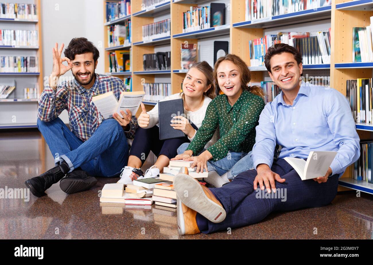 Eine Gruppe glücklicher Studenten, die zusammen in der Bibliothek studieren und auf dem Boden im Hintergrund mit Bücherregalen sitzen Stockfoto