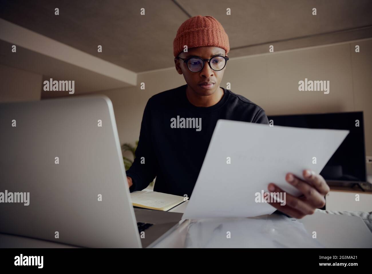 Afroamerikanischer Mann, der von zu Hause aus mit einem Laptop arbeitet. Glücklicher afroamerikanischer Mann, der Papier hält, während er von zu Hause aus hart arbeitet. Hochwertige Fotos Stockfoto