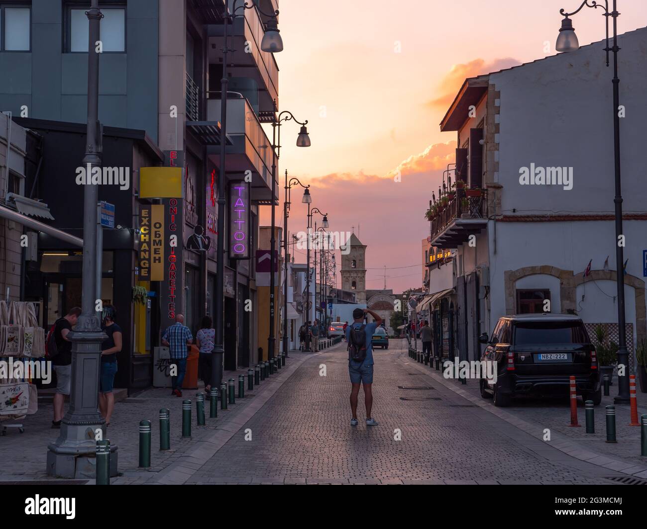 Ein Fotograf, der auf der Straße steht und ein Foto der lazarus Kirche in der Ferne mit einem farbenfrohen Abendhimmel bei Sonnenuntergang in Larnaca fotografiert. Stockfoto