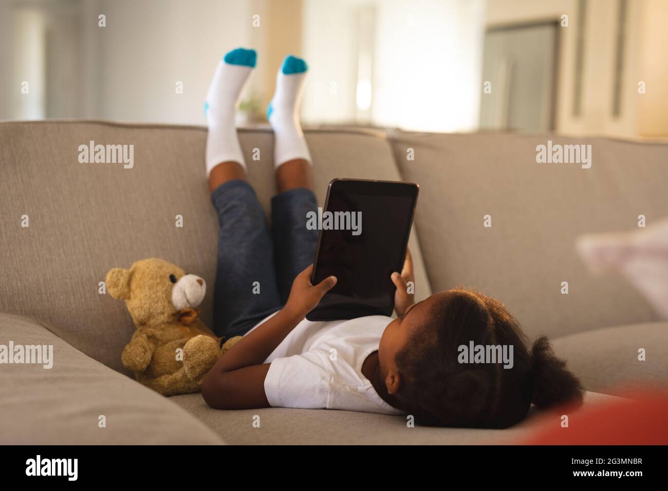 Glückliches afroamerikanisches Mädchen, das mit dem Tablet auf der Couch auf den Kopf gestellt wurde, kopierte Platz auf dem Bildschirm mit Teddybär Stockfoto