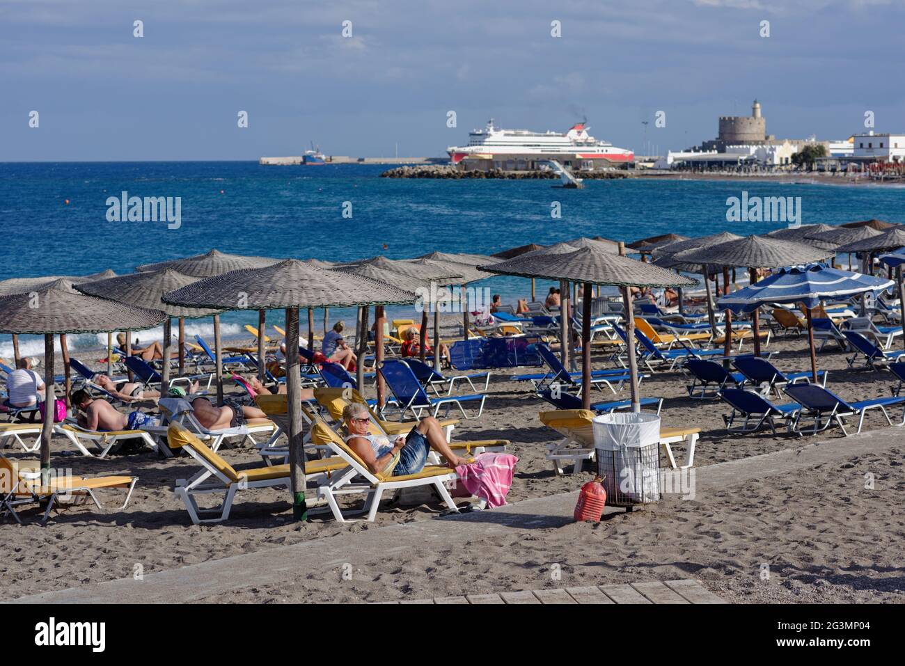 Menschen, die sich am Strand gegen die Bucht und den Hafen von Rhodos-Stadt, Rhodos-Insel, Dodekanes, Griechenland ausruhen Stockfoto